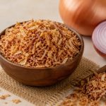 Prosesnya cukup sederhana, tetapi beberapa detail menentukan tekstur akhir. Bawang yang terlalu tebal sulit kering merata, sedangkan irisan terlalu tipis mudah berubah warna sebelum bagian dalamnya matang. Karena itu, siapkan alat, wadah peniris, dan tempat penyimpanan sebelum mulai menggoreng agar setiap tahap berjalan lancar. Cara Membuat Bawang Goreng Renyah 1. Pilih Bawang dengan Kondisi Baik Gunakan bawang merah yang masih segar, padat, tidak bertunas, dan bebas dari bagian busuk. Bawang dengan kadar air serta ukuran yang relatif seragam membantu irisan matang lebih merata saat digoreng. Kupas kulitnya, lalu buang bagian yang rusak sebelum mencuci bawang. Setelah mencuci, tiriskan dan keringkan permukaannya agar air tidak memicu percikan minyak ketika proses penggorengan dimulai. Langkah ini juga membantu minyak tetap stabil dan mengurangi risiko bawang saling menempel. 2. Iris dengan Ketebalan Seragam Iris bawang secara tipis dan konsisten supaya setiap potongan matang dalam waktu yang hampir sama. Pisau tajam dapat membantu pekerjaan skala kecil, sedangkan usaha dengan produksi lebih banyak bisa mempertimbangkan mesin perajang bawang dari Rumah Mesin agar proses pengirisan lebih cepat dan seragam. Jangan menumpuk irisan terlalu lama dalam keadaan basah karena kelembapan dapat membuat hasil gorengan kurang garing. Sebarkan irisan di atas wadah bersih dan biarkan permukaannya mengering sebelum masuk ke minyak. Jika menyiapkan banyak bawang, kerjakan secara bertahap agar irisan yang menunggu tidak menumpuk terlalu lama. 3. Gunakan Bumbu Secukupnya Sebagian orang menambahkan sedikit garam untuk memperkuat rasa bawang goreng. Jika memakai garam, campurkan tipis dan merata, lalu segera goreng agar bumbu tidak menarik terlalu banyak air dari irisan bawang. Pelaku usaha makanan juga bisa membuat satu uji coba tanpa tambahan bumbu untuk mengetahui rasa alami bawang. Hindari menambahkan banyak tepung jika ingin menghasilkan bawang goreng dengan tekstur ringan dan rasa bawang yang tetap menonjol. 4. Atur Suhu dan Jumlah Minyak Panaskan minyak dengan api sedang dan pastikan minyak cukup untuk membuat irisan terendam atau bergerak leluasa. Masukkan bawang dalam jumlah kecil agar suhu minyak tidak turun drastis dan irisan tidak saling menempel. Aduk perlahan selama menggoreng supaya panas menjangkau seluruh bagian. Saat bawang mulai berwarna kuning keemasan, segera angkat karena sisa panas masih dapat membuat warnanya semakin gelap setelah keluar dari penggorengan. Hindari meninggalkan wajan tanpa pengawasan karena perubahan warna dapat berlangsung cepat pada tahap akhir. 5. Tiriskan dan Dinginkan dengan Benar Angkat bawang sebelum warnanya menjadi cokelat tua, kemudian tiriskan menggunakan saringan atau kertas penyerap minyak khusus makanan. Sebarkan bawang di atas wadah lebar agar uap panas cepat keluar dan bagian bawahnya tidak menahan kelembapan. Jangan langsung memasukkan bawang yang masih panas ke dalam stoples. Tunggu sampai suhunya turun dan teksturnya kering, sebab uap yang terperangkap dapat membuat bawang cepat melempem. Pastikan wadah penyimpanan tidak menyimpan sisa air dari proses pencucian atau pemakaian sebelumnya. 6. Simpan dalam Wadah Kedap Udara Masukkan bawang goreng yang sudah dingin ke wadah kering dan kedap udara, lalu letakkan di tempat sejuk serta jauh dari sinar matahari. Gunakan sendok yang bersih dan kering setiap kali mengambilnya agar kelembapan tidak masuk ke dalam wadah. Untuk produksi rutin, buat catatan tanggal penggorengan dan jumlah hasil agar stok lama bisa digunakan lebih dulu. Informasi mengenai peralatan pengolahan pangan juga dapat ditemukan melalui situs Rumah Mesin sebagai referensi saat usaha membutuhkan proses kerja yang lebih efisien. Kesimpulan Cara membuat bawang goreng renyah bergantung pada kualitas bawang, ketebalan irisan, pengaturan suhu minyak, dan proses penirisan. Kerjakan setiap tahap dengan rapi agar bawang matang merata dan tidak menyerap minyak berlebihan. Setelah dingin, simpan hasil gorengan dalam wadah kedap udara untuk menjaga kerenyahannya. Jika membuat dalam jumlah besar, pertimbangkan alat perajang yang sesuai kapasitas supaya ukuran irisan lebih konsisten dan waktu persiapan lebih efisien.
